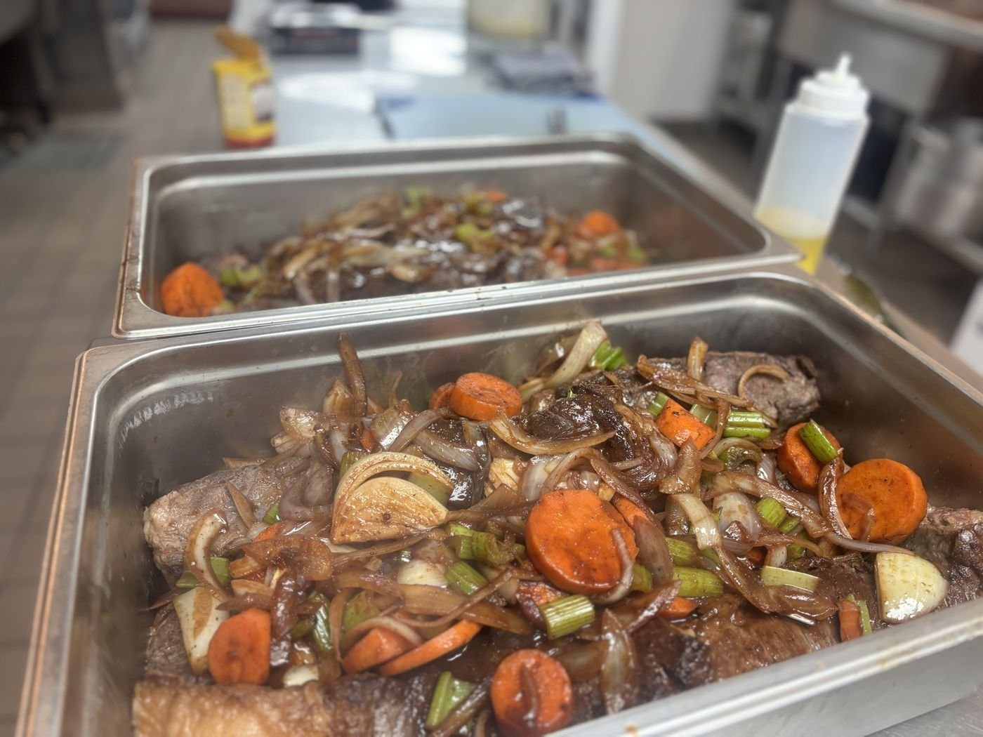 Two steam pans of braised beef with caramelized onions, carrots, and celery ready for service